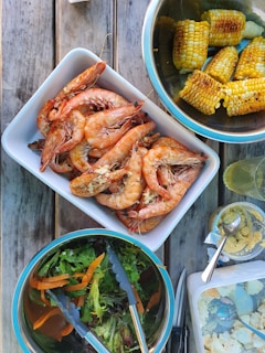 A wooden table is set with a variety of dishes featuring grilled prawns in a rectangular white dish, a bowl of corn on the cob, a mixed green salad with carrots in a bowl, and a container of potato salad. There is also a bowl of hummus with a spoon next to it and a glass of what appears to be a light drink.