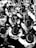 A large group of marching band members are playing instruments, all wearing uniforms with wide-brimmed hats and sporting logos on their jackets. The image is in black and white, creating a visually striking pattern with the uniformity and repetition of the figures and instruments.