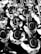 A large group of marching band members are playing instruments, all wearing uniforms with wide-brimmed hats and sporting logos on their jackets. The image is in black and white, creating a visually striking pattern with the uniformity and repetition of the figures and instruments.