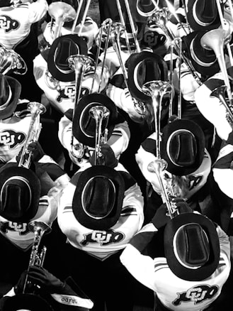 A large group of marching band members are playing instruments, all wearing uniforms with wide-brimmed hats and sporting logos on their jackets. The image is in black and white, creating a visually striking pattern with the uniformity and repetition of the figures and instruments.