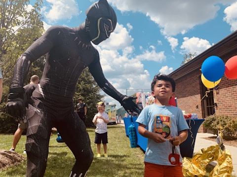 A child is standing outdoors on a grassy area, interacting with a person in a superhero costume. The sky is bright with fluffy clouds, and a building with colorful balloons attached is visible in the background. Other people and an inflatable structure are also in the scene.