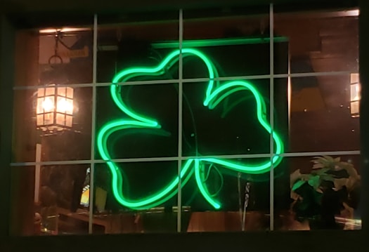 A neon green shamrock is glowing in the window of a dimly lit room or bar. The shamrock is framed by a window grid, and there is a softly lit lantern hanging on the left side. Some plants are visible at the bottom right corner.