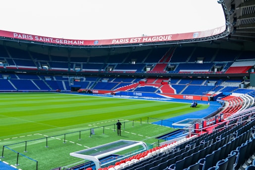 An empty football stadium with lush green grass on the field and blue and red seating areas. The stadium roof displays banners with writing, and the stands are well organized with alternating colors. A few maintenance vehicles and a person are visible near the pitch.