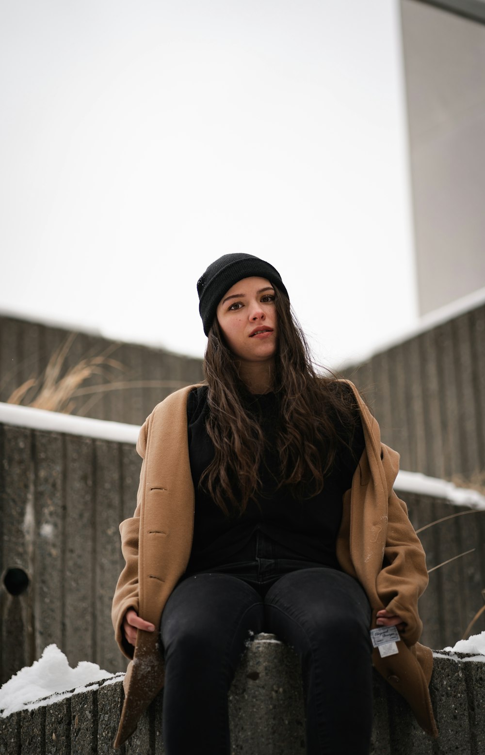 woman in brown coat and black pants sitting on white concrete wall during daytime