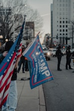 A street scene featuring individuals holding flags on the roadside. Prominent among them is a blue flag with red edges displaying the year 2020 and the phrase 'KEEP AMERICA GREAT.' American flags are also visible. Police officers are observed in the background, with tall buildings lining the street.
