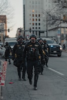 A group of law enforcement officers in tactical gear walk down a city street. Their uniforms include helmets, face masks, and protective vests. Behind them are police vehicles and a large, tall building. A leafless tree and city infrastructure are visible, alongside a sign with partial text.
