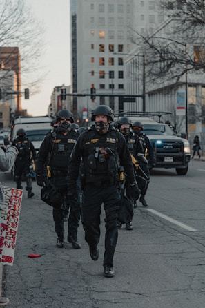 A group of law enforcement officers in tactical gear walk down a city street. Their uniforms include helmets, face masks, and protective vests. Behind them are police vehicles and a large, tall building. A leafless tree and city infrastructure are visible, alongside a sign with partial text.