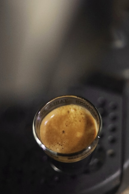A freshly brewed espresso in a small, transparent glass cup. The espresso has a rich, creamy crema on top, and it's placed on a dark, textured surface.