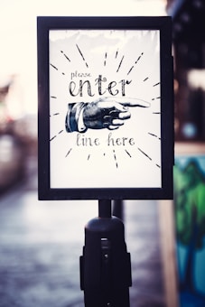 A sign with a vintage design featuring a hand illustration pointing to the right. The text reads 'please enter line here' in an ornate font, surrounded by decorative lines. The sign is encased in a black frame mounted on a pole.