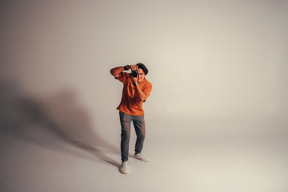 man in orange crew neck t-shirt and blue denim jeans standing and holding black camera