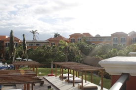 A charming resort setting with multiple orange-roofed buildings framed by lush greenery and tall palm trees. There are wooden pergolas in the foreground, providing a shaded area over patio seating. The sky is partly cloudy with a glare from the sun in the upper right corner.
