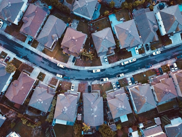 An aerial view of a suburban neighborhood with rows of houses lined along a curving street. The houses have similar architectural styles with gabled roofs, driveways, and small yards. Several cars are parked along the street and in driveways. The image captures a peaceful, residential area from a high angle.