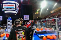 A group of people is engaged in a robotics competition environment. The scene includes a bright display screen showing match details, banners with competition logos, and scattered orange balls on the floor. Participants are focused on a piece of equipment or robot. The setting is within an indoor arena with seating for spectators.