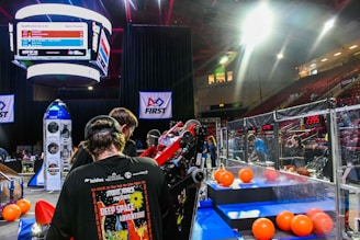 A group of people is engaged in a robotics competition environment. The scene includes a bright display screen showing match details, banners with competition logos, and scattered orange balls on the floor. Participants are focused on a piece of equipment or robot. The setting is within an indoor arena with seating for spectators.