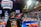 A group of people is engaged in a robotics competition environment. The scene includes a bright display screen showing match details, banners with competition logos, and scattered orange balls on the floor. Participants are focused on a piece of equipment or robot. The setting is within an indoor arena with seating for spectators.