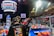 A group of people is engaged in a robotics competition environment. The scene includes a bright display screen showing match details, banners with competition logos, and scattered orange balls on the floor. Participants are focused on a piece of equipment or robot. The setting is within an indoor arena with seating for spectators.