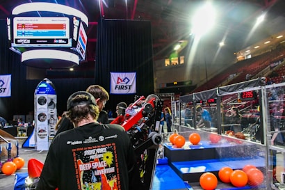 A group of people is engaged in a robotics competition environment. The scene includes a bright display screen showing match details, banners with competition logos, and scattered orange balls on the floor. Participants are focused on a piece of equipment or robot. The setting is within an indoor arena with seating for spectators.