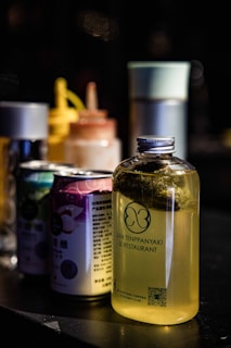 A set of containers is displayed on a dark surface, including a clear bottle filled with a yellow liquid and labeled 'San Tenppanyaki & Restaurant'. Behind the bottle are a variety of colorful cans and bottles, some with spray nozzles, arranged in a line creating a visually appealing arrangement.