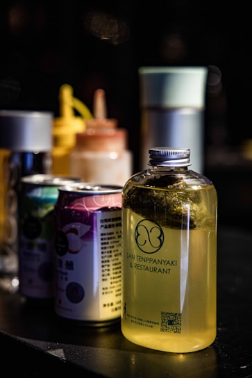 A set of containers is displayed on a dark surface, including a clear bottle filled with a yellow liquid and labeled 'San Tenppanyaki & Restaurant'. Behind the bottle are a variety of colorful cans and bottles, some with spray nozzles, arranged in a line creating a visually appealing arrangement.
