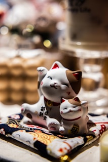 Two ceramic cat figurines are placed on a colorful patterned fabric. The larger cat has its paw raised and features a welcoming smile with a collar around its neck. The smaller cat sits closely beside the larger one, mirroring its smile.