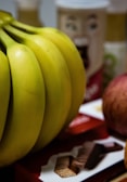 A close-up view of a bunch of ripe bananas with their smooth yellow skins. In the background, there's a red and white package featuring chocolate wafers, an apple, and a container with an illustrated face.