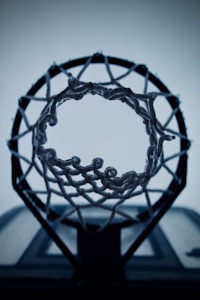A basketball hoop is viewed from below, showcasing the net's intricate weaving against a pale, blurred background. The focus on the hoop's details creates a dramatic and intense perspective.