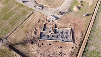 An aerial view of a construction site where the foundation of a building is being prepared. The image shows a rectangular structure with wooden framing and steel reinforcements embedded in concrete. Surrounding the construction area are patches of bare earth and some green grass. Construction vehicles, including a bulldozer and an excavator, are positioned near the site.