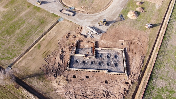 An aerial view of a construction site where the foundation of a building is being prepared. The image shows a rectangular structure with wooden framing and steel reinforcements embedded in concrete. Surrounding the construction area are patches of bare earth and some green grass. Construction vehicles, including a bulldozer and an excavator, are positioned near the site.