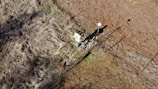A person wearing a yellow vest stands near a fence on uneven, grassy terrain. They appear to be using a surveying instrument mounted on a red and white pole. The image is captured from a high angle, showing the surrounding dry grass and dirt patches.