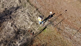 A person wearing a yellow vest stands near a fence on uneven, grassy terrain. They appear to be using a surveying instrument mounted on a red and white pole. The image is captured from a high angle, showing the surrounding dry grass and dirt patches.
