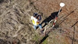 A person wearing a reflective safety vest stands outdoors on grassy terrain. They are holding equipment and seem engaged in some form of land survey work. Nearby, a tripod with a white circular device is set up, casting a shadow on the ground.