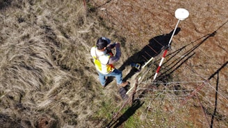 A person wearing a reflective safety vest stands outdoors on grassy terrain. They are holding equipment and seem engaged in some form of land survey work. Nearby, a tripod with a white circular device is set up, casting a shadow on the ground.