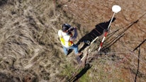 A person wearing a reflective safety vest stands outdoors on grassy terrain. They are holding equipment and seem engaged in some form of land survey work. Nearby, a tripod with a white circular device is set up, casting a shadow on the ground.