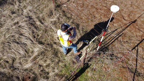 A person wearing a reflective safety vest stands outdoors on grassy terrain. They are holding equipment and seem engaged in some form of land survey work. Nearby, a tripod with a white circular device is set up, casting a shadow on the ground.