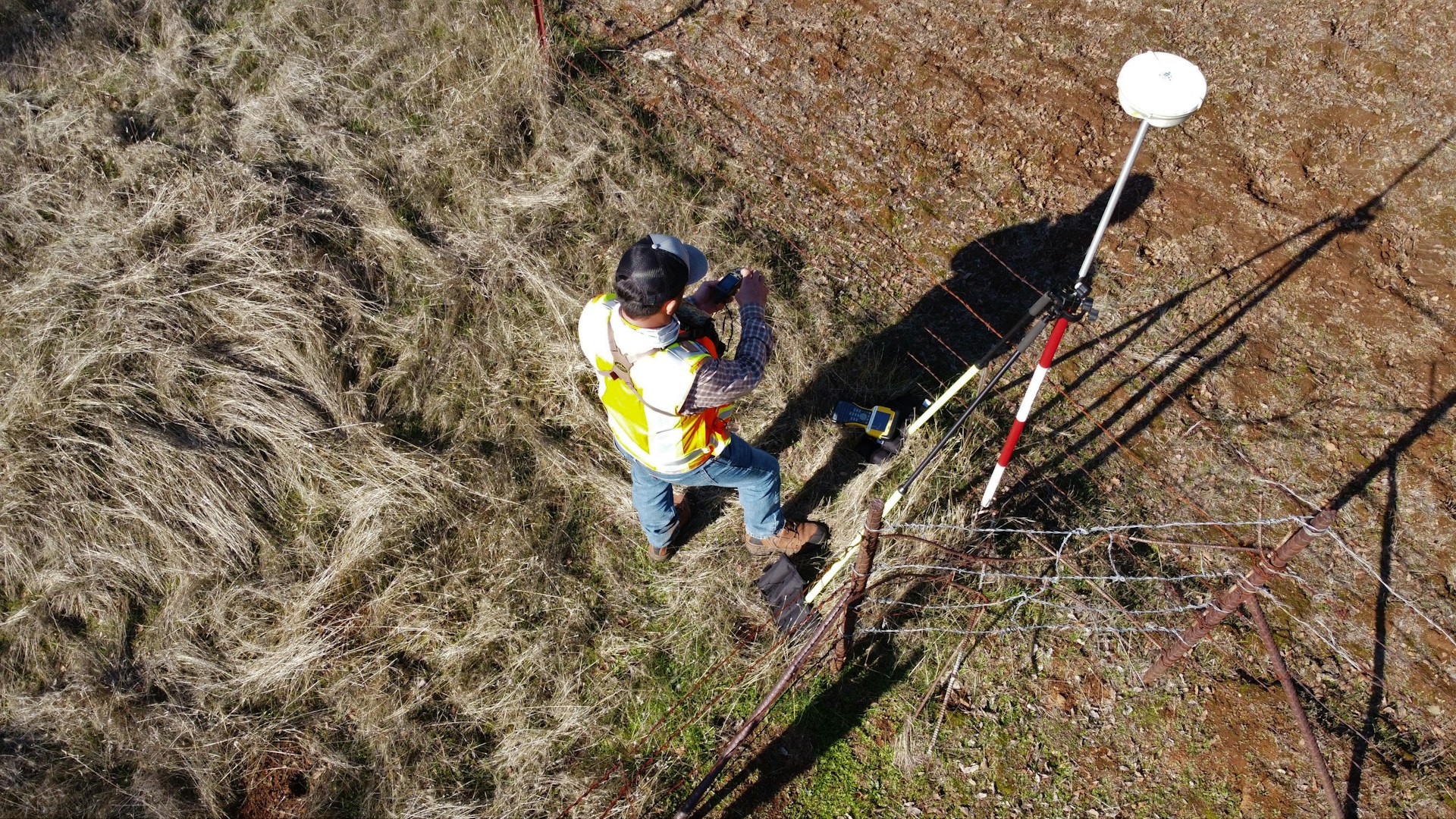A person wearing a reflective safety vest stands outdoors on grassy terrain. They are holding equipment and seem engaged in some form of land survey work. Nearby, a tripod with a white circular device is set up, casting a shadow on the ground.