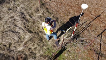 A person wearing a reflective safety vest stands outdoors on grassy terrain. They are holding equipment and seem engaged in some form of land survey work. Nearby, a tripod with a white circular device is set up, casting a shadow on the ground.