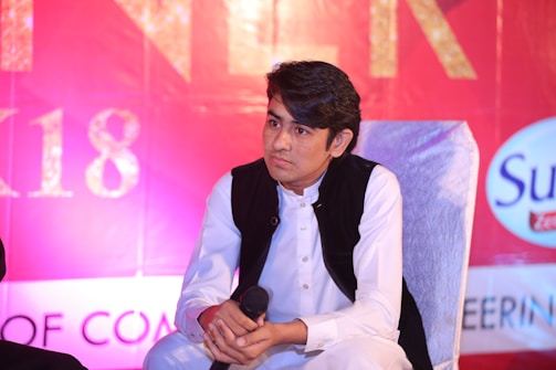A man with dark hair is seated on a chair, wearing a white traditional outfit with a black vest. He is holding a microphone and appears to be listening attentively. The background is predominantly red with some text partially visible, including the letters '18' and a logo on the right-hand side.