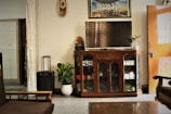 A living room interior with a focus on a wooden cabinet featuring glass doors. On top of the cabinet is a flat-screen TV and a small decorative plant. An artwork hangs above the TV. A potted plant sits to the left of the cabinet beside a small speaker. The room includes a sofa with visible armrests, a partially open wooden door on the right, and a curtain separating one area from another. A decorative clock is mounted on the wall.