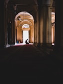 A person is kneeling in a dimly lit corridor with tall columns on either side. The light is coming from a large arched doorway with stained glass panels at the top. The setting suggests a tranquil and reflective atmosphere.