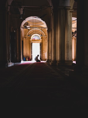 A person is kneeling in a dimly lit corridor with tall columns on either side. The light is coming from a large arched doorway with stained glass panels at the top. The setting suggests a tranquil and reflective atmosphere.