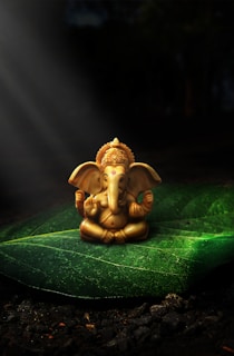 A small, intricately detailed statue of a deity with an elephant head rests on a large, verdant leaf. Sunlight streams down, illuminating the statue, creating a serene and spiritual atmosphere. The background is dark and blurred, drawing focus to the statue and leaf.