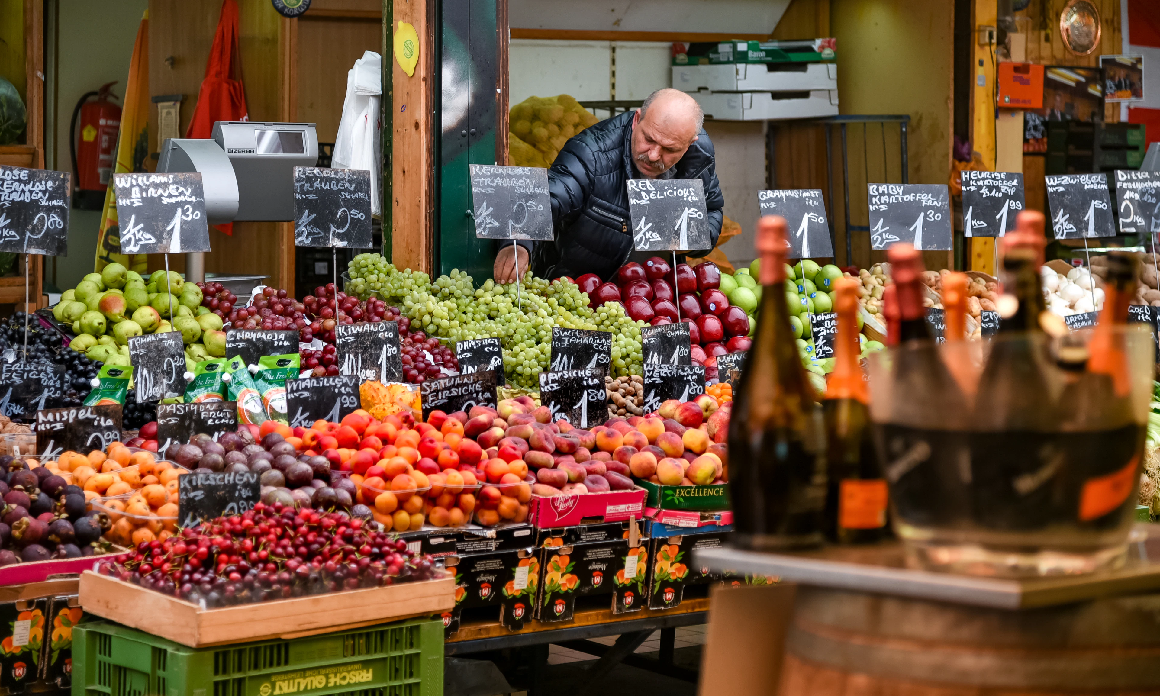 A fruit and vegetable stall at Vienna's Naschmarkt, its crates heaped with grapes, apples, tomatoes and stone fruit under handwritten chalkboard price tags, a bottle display in the foreground and the stallholder at work behind the counter.