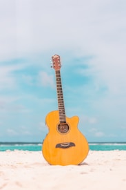 A wooden acoustic guitar stands upright on a pristine beach with fine white sand. The background features a serene blue sky and calm turquoise ocean waves in the distance.