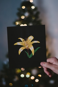 A hand holds a black card featuring a yellow lily with green leaves. In the background, there are blurred lights resembling a softly focused Christmas tree adorned with yellow and white bokeh lights.