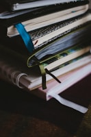 A stack of notebooks or journals with different colored covers and ribbon bookmarks is piled on a dark surface. The covers are textured, varying in patterns and colors, and the pages appear to be slightly used, suggesting regular use.