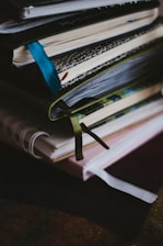 A stack of notebooks or journals with different colored covers and ribbon bookmarks is piled on a dark surface. The covers are textured, varying in patterns and colors, and the pages appear to be slightly used, suggesting regular use.