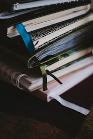 A stack of notebooks or journals with different colored covers and ribbon bookmarks is piled on a dark surface. The covers are textured, varying in patterns and colors, and the pages appear to be slightly used, suggesting regular use.