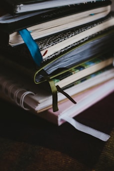 A stack of notebooks or journals with different colored covers and ribbon bookmarks is piled on a dark surface. The covers are textured, varying in patterns and colors, and the pages appear to be slightly used, suggesting regular use.