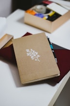A journal with a simple design of a white branch and leaves on a brown cover rests on a table. Next to it, a box of stationery supplies, including tape and colored circles, is visible. A pen is tucked into the journal, which sits on top of a closed burgundy folder.