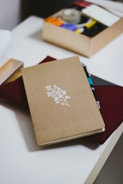 A journal with a simple design of a white branch and leaves on a brown cover rests on a table. Next to it, a box of stationery supplies, including tape and colored circles, is visible. A pen is tucked into the journal, which sits on top of a closed burgundy folder.